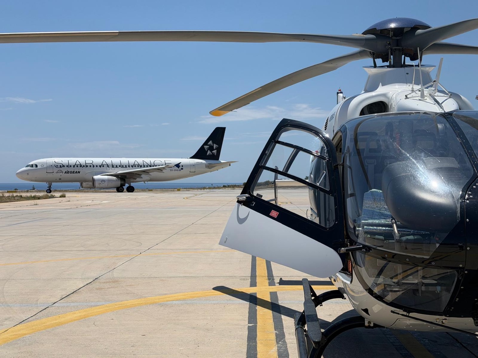 Fly G Aviation Airbus H135 helicopter at Santorini Airport with Star Alliance Aegean Airlines aircraft in the background during summer operations in Greece