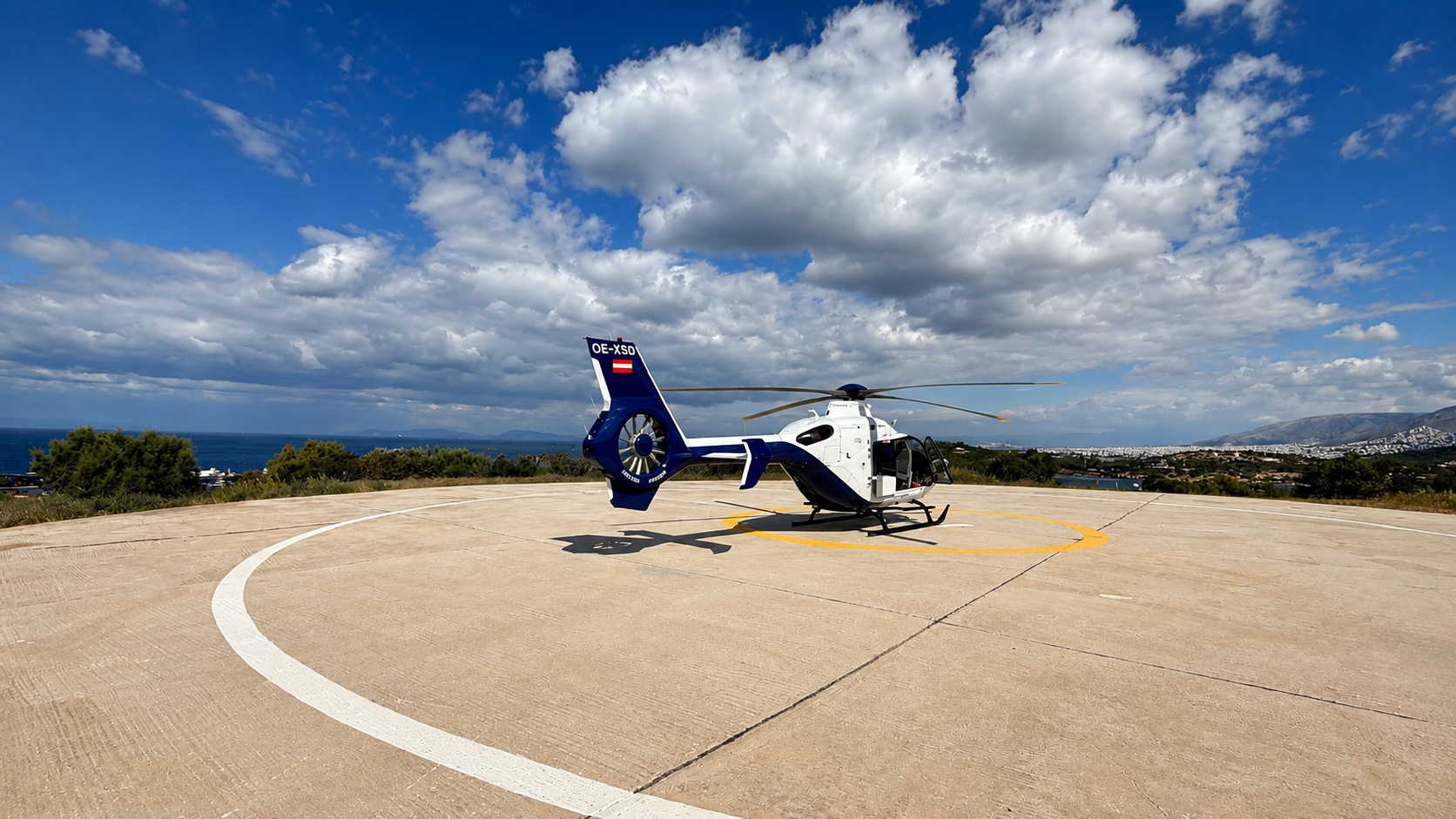 Airbus H135 helicopter at the Four Seasons Athens helipad overlooking the Aegean coastline under dramatic clouds